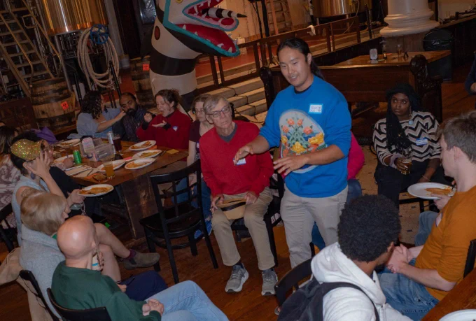 Community organizer Alex Zhu stands and speaks to a diverse group of people gathered around wooden tables inside a spacious brewery or event hall. Zhu is wearing a bright blue sweatshirt with a colorful graphic and a nametag, gesturing with his hands as the seated attendees look on.