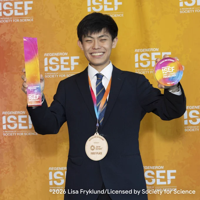 Regeneron ISEF 2026 において、満面の笑みで写る栗林輝さん。濃紺のスーツを着用し、オレンジ色のイベント背景幕を背にしている。右手にはカラフルなグラデーションのトロフィー、左手には同じグラデーションの丸い大会公式メダルを掲げ、首からはスポンサー企業から贈られた大きな木製メダルが掛けられている。