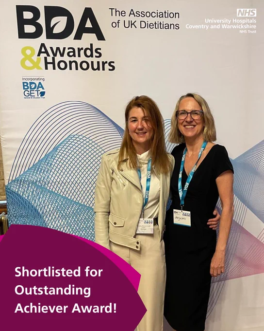 Two people stand side by side at the BDA Awards and Honours event in front of a branded backdrop reading “The Association of UK Dietitians.” Both are wearing event lanyards and name badges. One person is dressed in a light-coloured outfit and the other in a black dress. A graphic in the bottom left corner reads, “Shortlisted for Outstanding Achiever Award!” The NHS University Hospitals Coventry and Warwickshire logo appears in the top right corner.