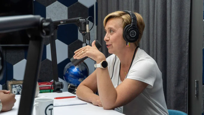 A woman wearing headphones speaks into a microphone in a recording studio, with a notebook and coffee mug on the table in front of her.