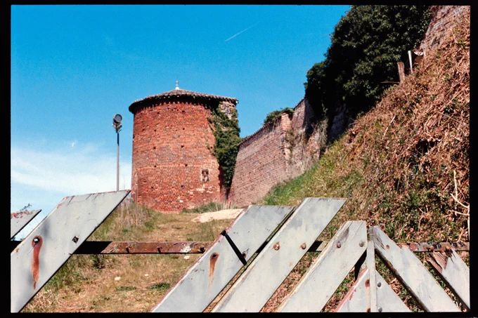 a picture of an old stone tower, made with red bricks, at the end of a little grassy path. from the tower goes a red brick wall as well, and in the foreground is a metal-and-painted-wood fence that's kind of falling apart but clearly blocks the path