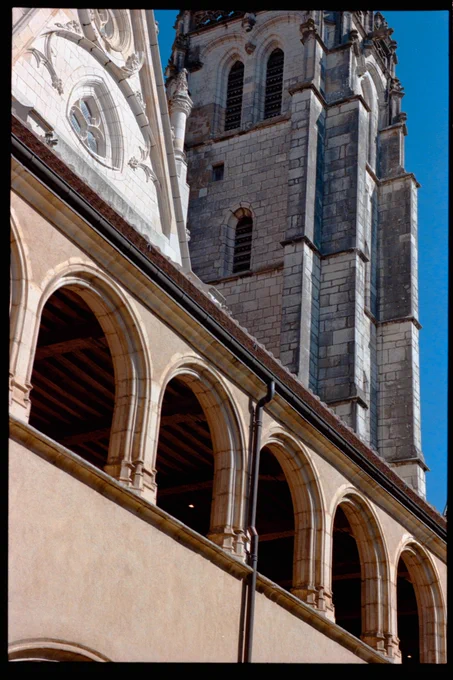 details of a large cathedral building. The lower level is a brown-yellow smooth material, with large arched openings along the top, then above that is a traditional church whitestone roof with a couple stained glass windows in it, and in the background is a much higher tower also made of white stone and very church-y.