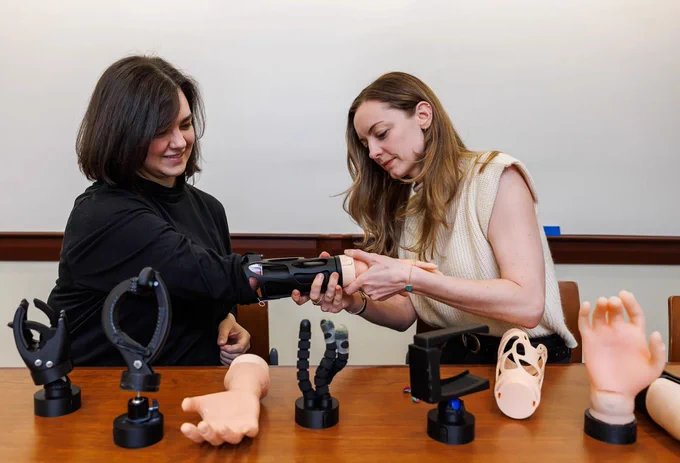 Two individuals are at a table displaying prosthetic arms and hands. They are focused on adjusting a prosthetic arm. The table holds various prosthetic designs and models.