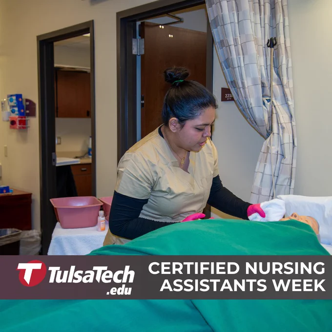 Nursing assistant wearing gloves providing bedside care to a patient covered with a green blanket in a clinical room.