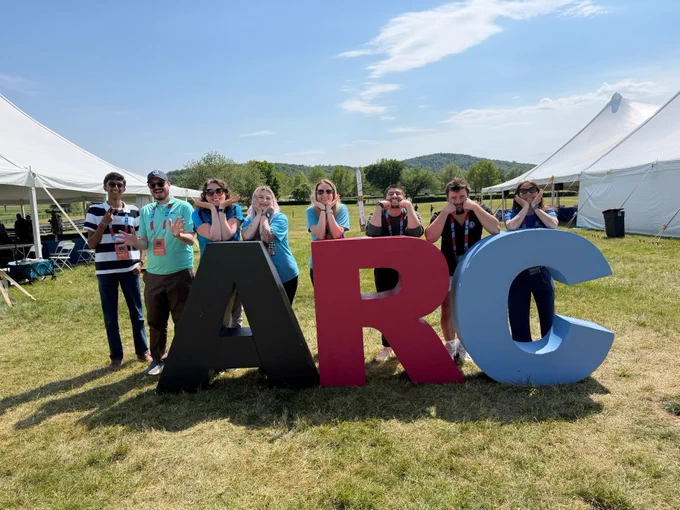 A group of eight people posing outdoors on a sunny day behind large, colorful, three-dimensional letters that spell "ARC" at the American Rocketry Challenge.