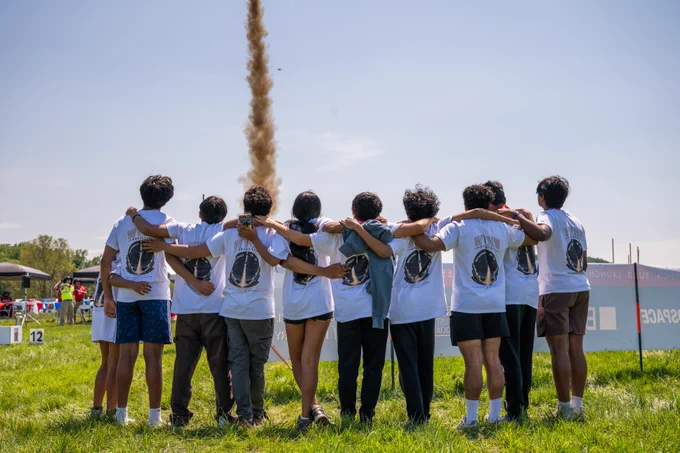A group of students with their backs to the camera wear matching team T-shirts. They stand in a grassy field, looking up at the sky as their model rocket ascends, leaving a trail of smoke.