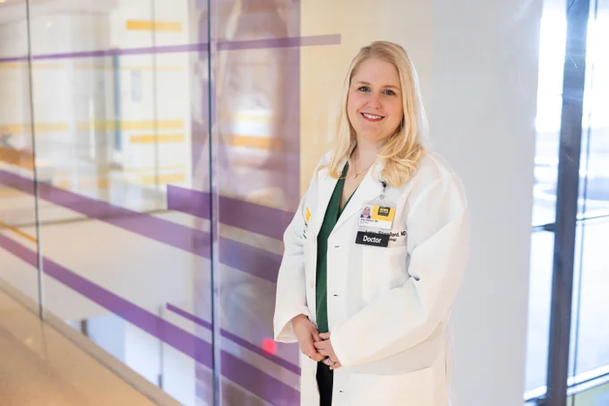 A woman with long blonde hair, wearing a white lab coat with a name tag that reads "Doctor," stands in a brightly lit hallway. The background features glass panels with purple and yellow stripes.