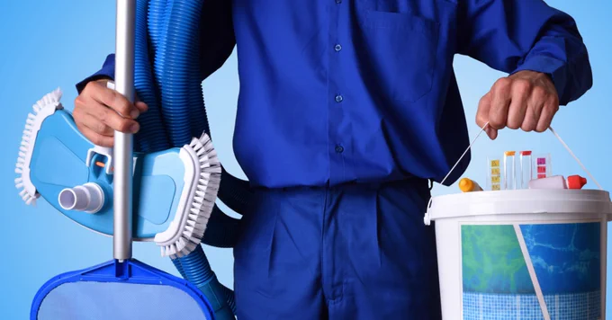 A person in blue overalls holds pool cleaning equipment, including a vacuum hose and a bucket with test strips and cleaning supplies.