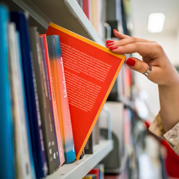 A hand with red nail polish selects an orange book from a library shelf.