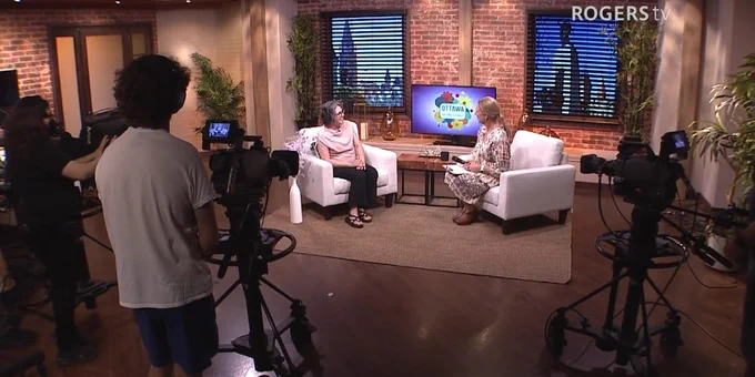 Two women seated in a cozy TV studio set with brick walls, engaged in conversation while cameras record them for Rogers TV.