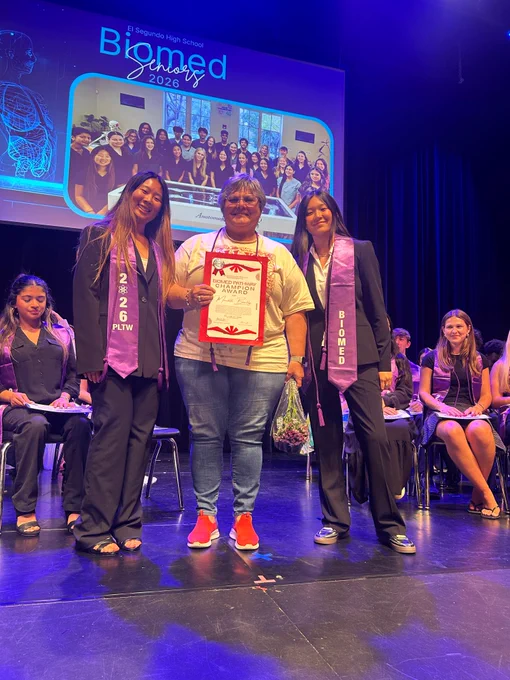 Three individuals stand on stage during a Biomed graduation ceremony, one holding a certificate and flowers, with a group photo projected behind them.