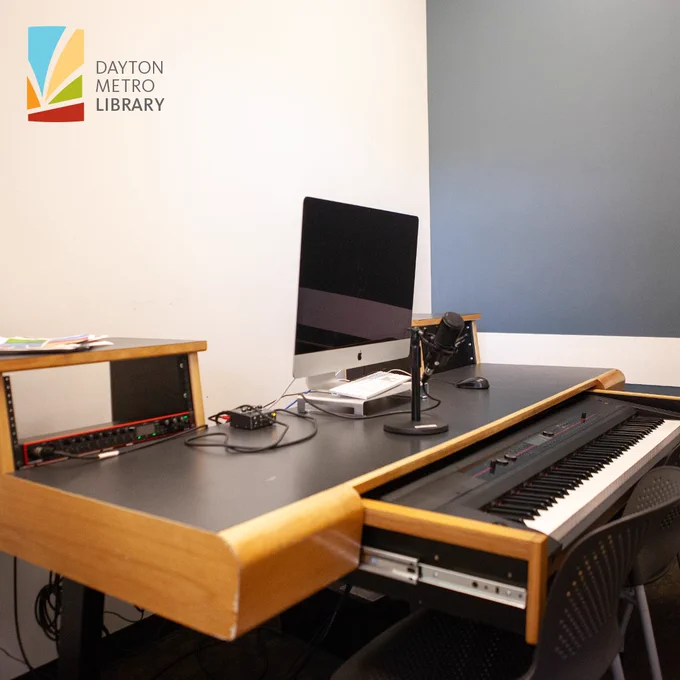 Recording desk with a desktop computer, microphone on a stand, and a slide-out digital piano keyboard in a Dayton Metro Library tech studio.