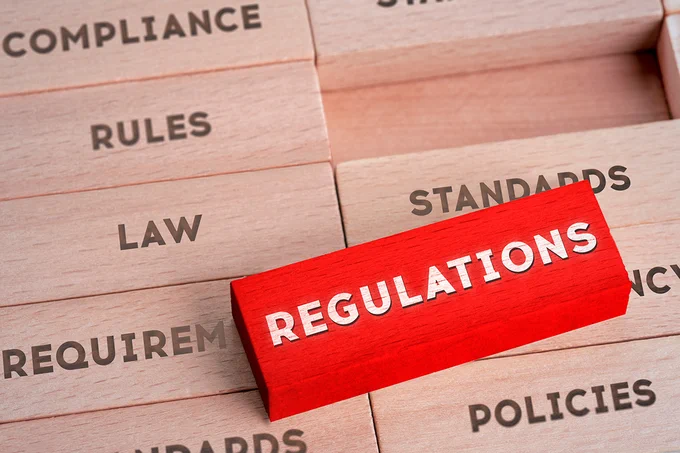 Red block labeled 'REGULATIONS' among wooden blocks with words like 'COMPLIANCE', 'RULES', 'LAW', and 'POLICIES'.
