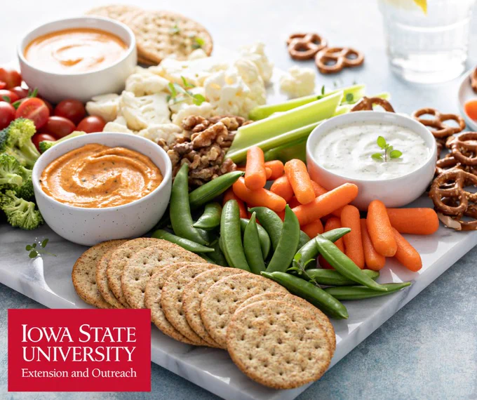 Assorted fresh vegetables, fruits, crackers, pretzels, and two creamy dips arranged on a white serving board.