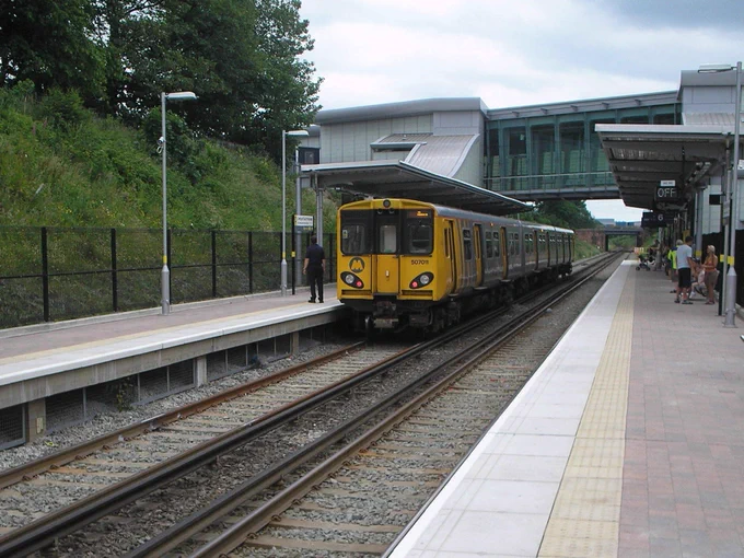 Merseyrail unit 507011 in "tiger stripe" livery calls at the Hunts Cross-bound platform at Liverpool South Parkway. There are passengers waiting on the opposite platform for their train.