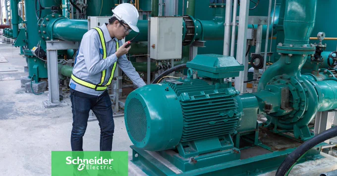 Eine Person mit Schutzhelm und Warnweste, die ein Walkie-Talkie in der Hand h\u00E4lt und eine gro\u00DFe, blaugr\u00FCne Industriepumpe sowie Rohrleitungen inspiziert. 