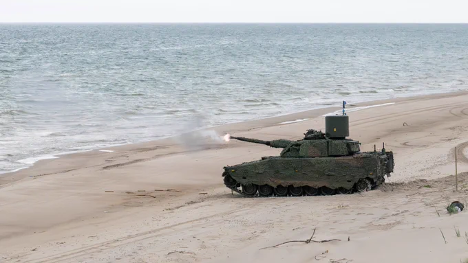 Military tank firing a shell on a sandy beach near the ocean shore under a clear sky.