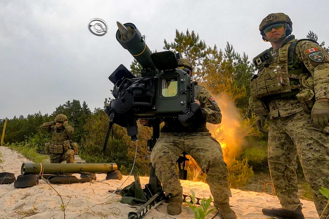 Soldiers in camouflage gear firing an anti-tank missile launcher on a sandy coastal area with trees in the background.