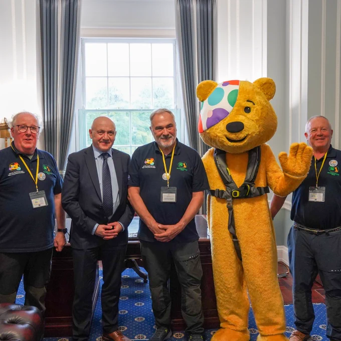 Pudsey Bear stands inside a bright office, wearing a safety harness and raising one paw, alongside four adults. One person is in formal business attire (The Assembly Deputy Speaker)while the others wear matching charity polo shirts with lanyards, standing in front of a large window with curtains and a desk behind them.