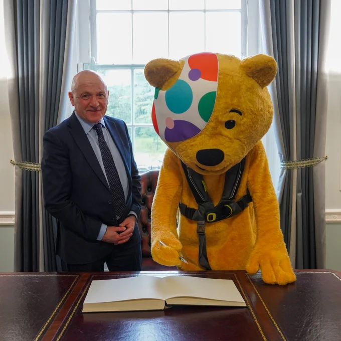 
Pudsey Bear stands at a wooden desk inside a formal office, writing in an open visitors’ book, while a man wearing a suit and tie, the Assembly Deputy Speaker John Blair MLA, stands beside Pudsey. Tall windows with curtains and decorative gold stands are visible in the background.
