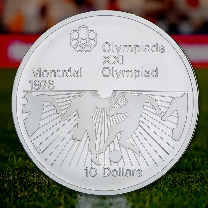 Pièce en argent commémorant les Jeux olympiques de 1976 à Montréal. On y voit les anneaux olympiques et les silhouettes d’athlètes en mouvement, dont une qui botte un ballon, et les inscriptions « Montréal 1976 », « Olympiade XXI Olympiad » et « 10 Dollars ».

