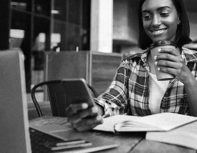 Person in a plaid shirt holding a cup and looking at a smartphone while sitting at a table with an open notebook and laptop.