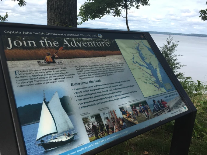 Informational sign at Captain John Smith Chesapeake National Historic Trail with a sailing ship image and a map of the trail along the water's edge.