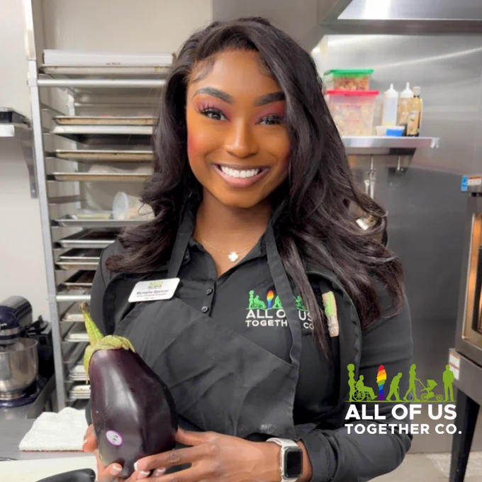 An image featuring a smiling, standing woman in cooking apron, holding an eggplant. Company logo positioned right in footer.
