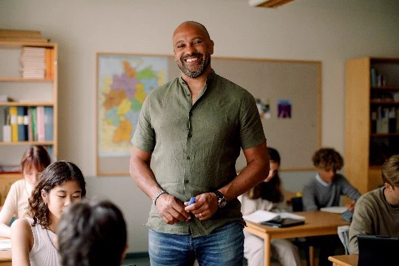 A smiling teacher stands at the front of a classroom while students work at their desks in the background.