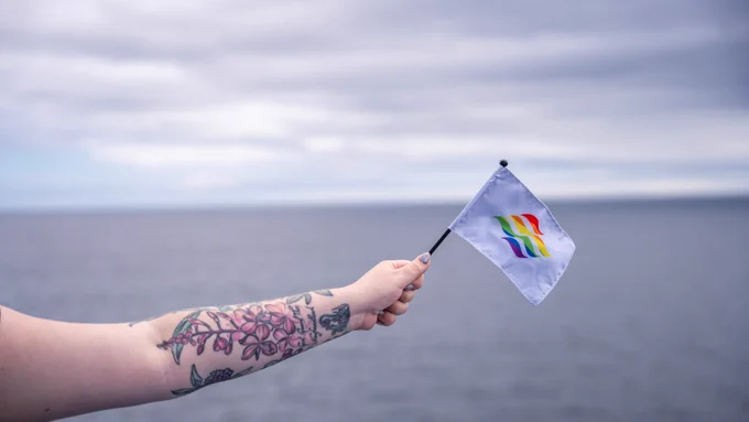 Photo shows arm with floral tattoos holding a flag with the Marine Atlantic logo in pride colours. Background is the ocean, with calm, blue waters. The sky is cloudy but bright. 