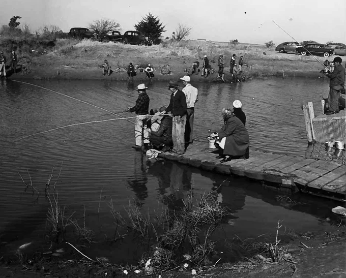 Historic black-and-white photograph showing a large crowd gathered along the shoreline of a lake or reservoir. People stand, sit, and walk near the water\u2019s edge, while others are scattered across the grassy bank. Several small boats can be seen on the water in the background, and rolling hills or open countryside stretch beyond the lake. The image depicts a busy outdoor recreation scene from an earlier era.