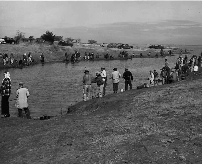 Historic black-and-white photograph showing a large crowd gathered along the shoreline of a lake or reservoir. People stand, sit, and walk near the water\u2019s edge, while others are scattered across the grassy bank. Several small boats can be seen on the water in the background, and rolling hills or open countryside stretch beyond the lake. The image depicts a busy outdoor recreation scene from an earlier era.