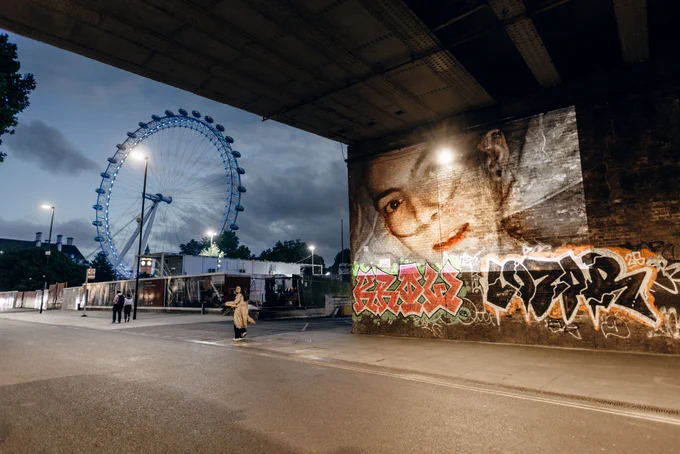 A woman's image, lying in bed with a breathing tube is projected under a bridge with the London Eye in the background.