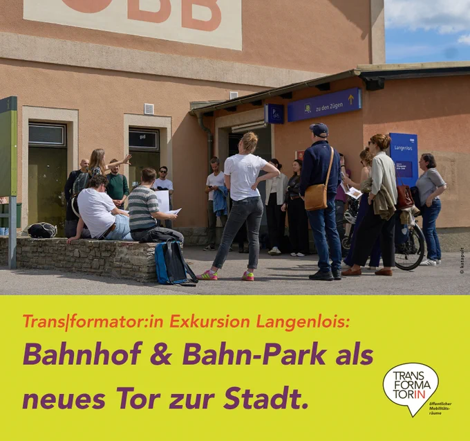 Eine Menschengruppe steht bzw. sitzt (auf Steinsockel) am Vorplatz vor einem orangen Gebäude (dem Bahnhofsgebäude) und hört einer vortragenden Frau zu, die auf etwas deutet.