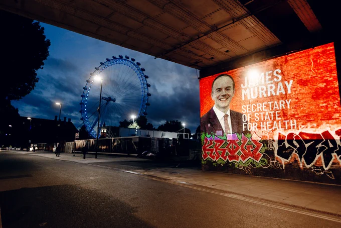 A photograph of James Murray with white text on red background -  'James Murray Secretary of State for Health' in white  is projected under a bridge with the London Eye in the background.