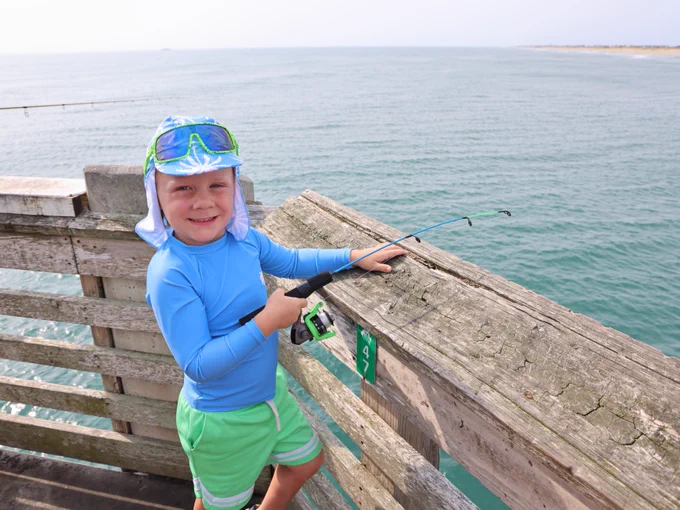 A boy wearing a sun hat and blue sun-protective clothing is smiling while holding a fishing rod on a wooden pier railing overlooking the ocean. The scene appears calm and sunny.