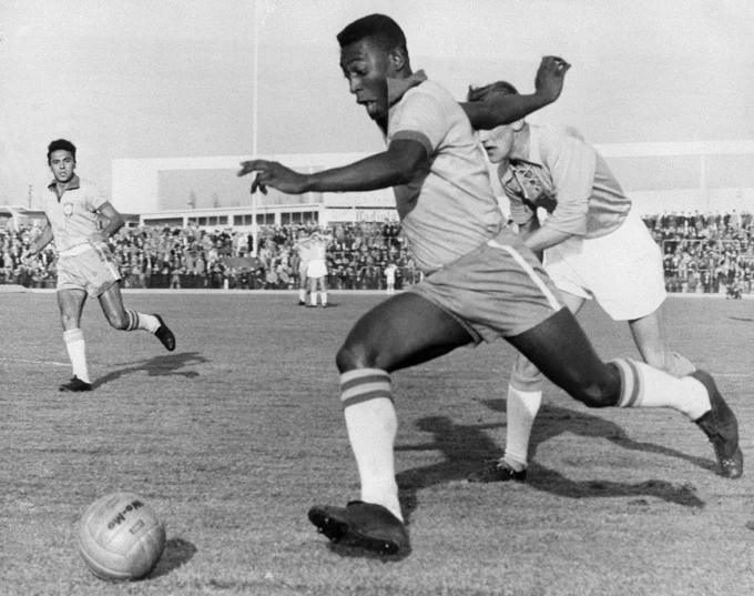 A black-and-white photo shows Pelé dribbling  a soccer ball past several opposing team members, while stands full of onlookers watch.