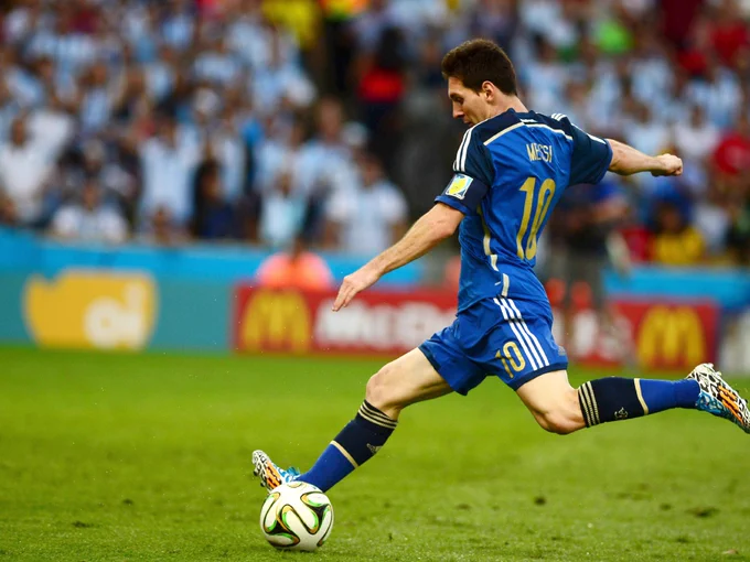 Messi, in blue jersey and shorts, prepares to kick the ball on a grass field, crowd blurred in background.