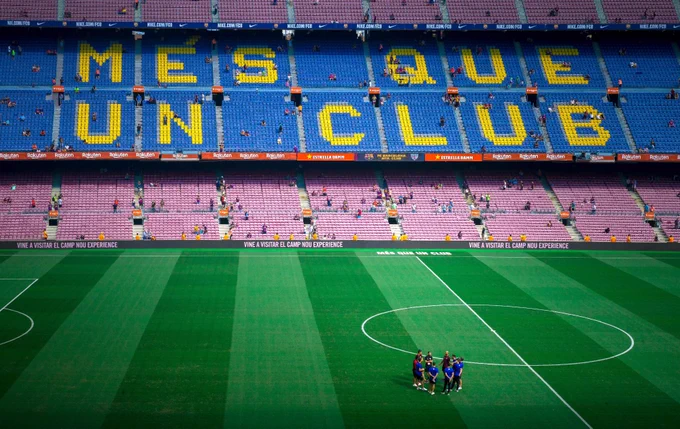 Players circle up in the field at a soccer stadium, while a smattering of fans watches on from the stadium seats. The soccer club’s motto is painted in yellow on a blue row of seats, reading “Més que un club.”