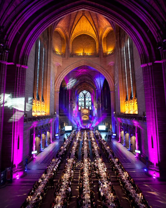 Long rows of dining tables arranged down the centre of a large cathedral nave, decorated for an event with candles and place settings, under dramatic purple and amber lighting highlighting the high vaulted ceiling and distant altar.