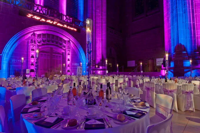 Round banquet tables set with white linens, glassware, wine bottles, and tall candle centrepieces fill a large cathedral interior, illuminated by vivid purple, pink, and blue lighting that highlights gothic stone arches and columns, with a neon-style sign glowing above an ornate doorway in the background.