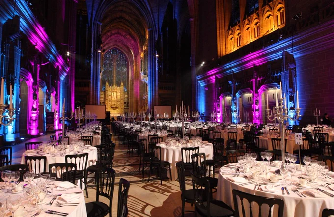 Round banquet tables set with white tablecloths, glassware, and silver candelabras fill a grand cathedral hall, illuminated by vibrant purple, blue, and warm amber lighting against tall gothic arches and stone walls.