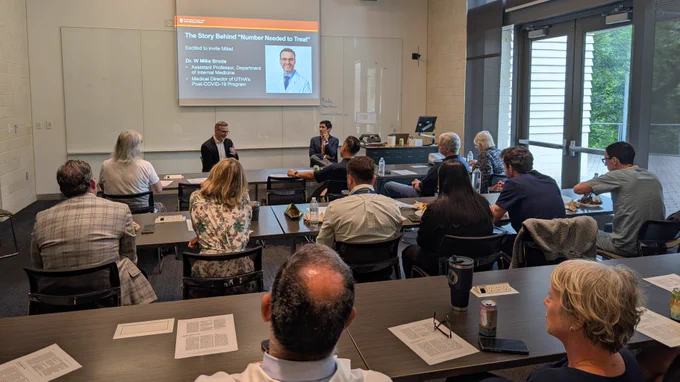 A group of people are seated in a conference room attending a presentation. Two individuals are seated at the front, engaging with the audience.