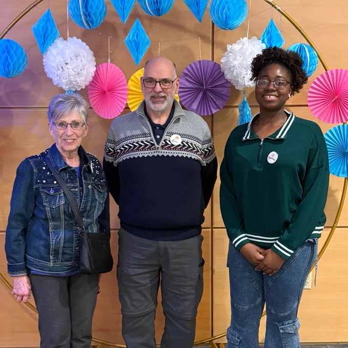 Three people pose together in front of a circular decorative backdrop with colourful hanging paper decorations. An older woman in a denim jacket, a man in a patterned sweater, and a younger woman in a green sweater all smile toward the camera.
