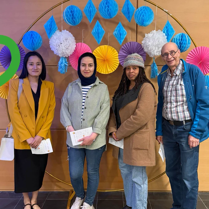 Four people stand together smiling in front of a circular decorative backdrop with hanging blue, pink, yellow, and purple paper decorations. Each person is holding a paper activity sheet at what appears to be a volunteer appreciation or community event.