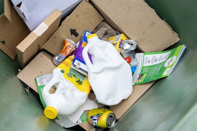 A green recycling bin overflowing with cardboard, crushed cans, plastic jugs, and food packaging.