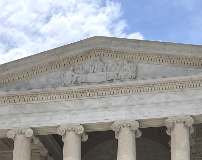 Close-up of the marble facade of the Jefferson Memorial with the tops of columns and a detailed sculpted frieze showing five men around a table.