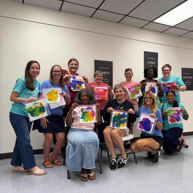 Group of ten people posing indoors, each holding colorful abstract paintings on paper, smiling and seated or standing against a white wall with motivational posters.