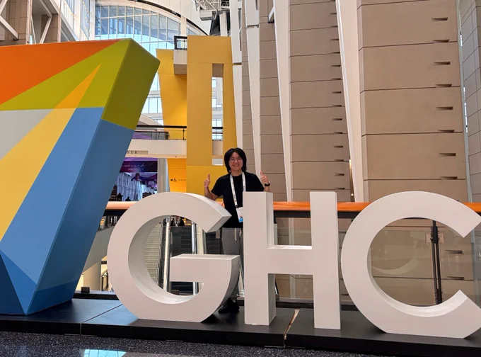 Student standing behind large white letters spelling 'GHC' inside a modern building with colorful geometric decor and tall windows.