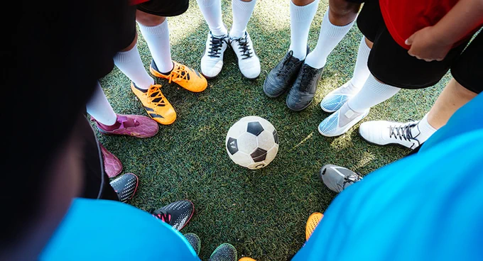 A group of kids wearing colorful cleats and white socks gathered around a soccer ball on grass.